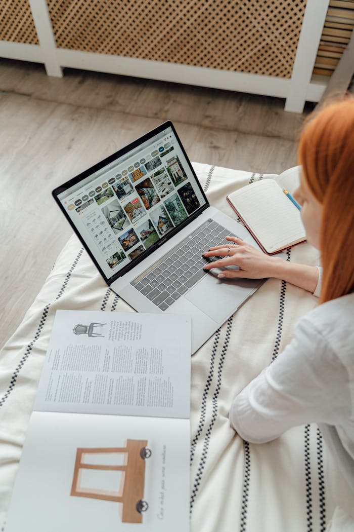 A woman comfortably works on a laptop in a cozy bedroom, surrounded by books and notes.